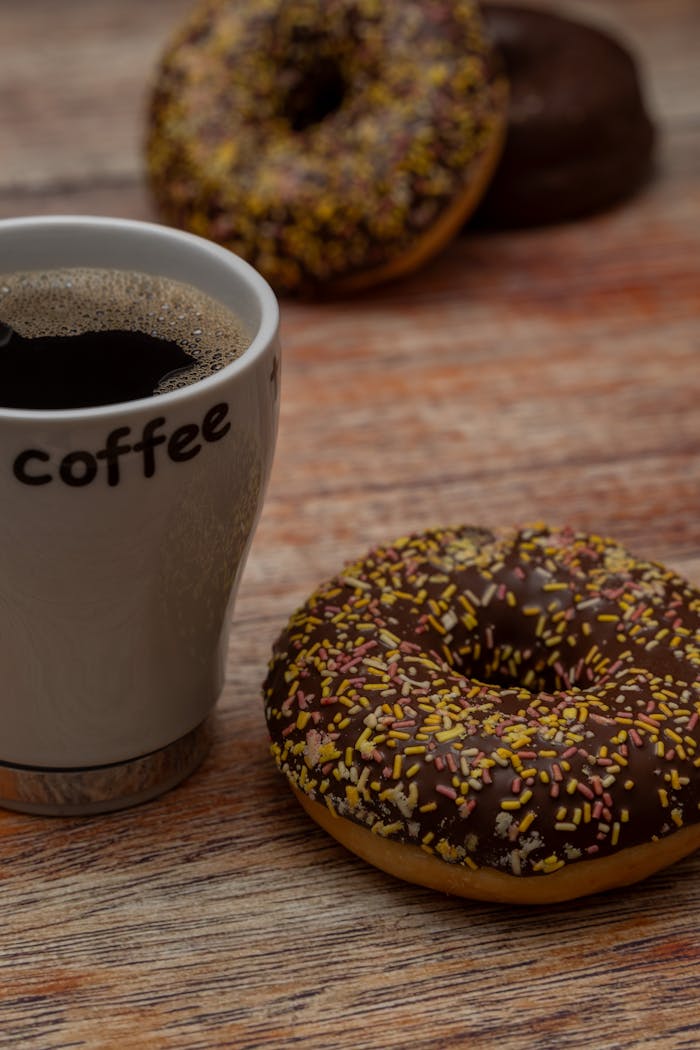 Delicious chocolate donuts with sprinkles and a hot cup of coffee on a rustic wood surface, perfect for breakfast or snack time.